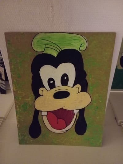 A painting of the cartoon character Goofy is displayed on a flat surface. The painting is framed and depicts Goofy's head and upper torso against a mottled green and gold background. Goofy is wearing a green hat and has a large, open mouth with a red tongue. His eyes are large and white with black pupils. The painting is lit from above. The background is a white wall with a textured surface. To the left of the painting, a white object with horizontal lines is partially visible, and to the right, a white object with a rectangular shape and a slight tilt is seen. The setting is indoors. This image was taken in Anderlecht, Belgium.