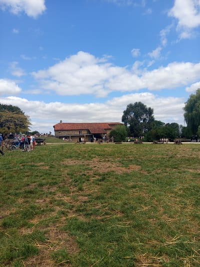 The image depicts an outdoor scene at a historical reenactment or museum in Azannes-et-Soumazannes, France. A large, traditional building with a red tile roof and stone walls is visible in the background, situated on a gentle slope. Several people, including adults and children, are gathered around the building and in the surrounding grassy area. Some are observing exhibits or demonstrations, while others appear to be casually interacting. In the foreground, a mowed grassy field with patches of dry straw is present. Various old farm implements and carts are scattered across the landscape, suggesting a historical farming context. The sky is a bright blue with scattered white clouds. The overall impression is of a sunny day at a public event focused on historical preservation and education.