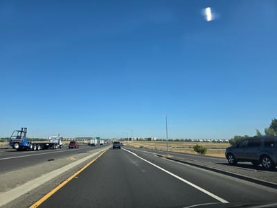The image depicts a clear, sunny day with a deep blue sky. The perspective is from inside a vehicle traveling on a highway, likely in or near Sacramento, California, based on the vegetation and urban sprawl visible in the distance.
The highway has multiple lanes and is paved with asphalt. White and yellow lane markers are visible. To the left, a large blue forklift is being transported on a flatbed truck. Other vehicles, including cars and trucks, are also on the highway.
The environment includes dry, grassy fields on either side of the highway, with a line of trees and distant buildings that suggest a suburban or exurban area. A streetlamp stands tall on the right side of the road. The overall scene is one of ordinary highway travel on a bright day.