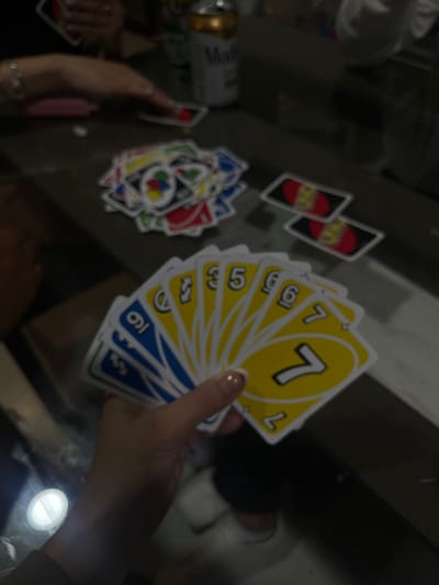 A slightly blurred image shows a hand holding several Uno cards, the top one being a yellow 7. A pile of discarded Uno cards sits on the table. Two red "Uno" cards are also visible nearby. A can of Modelo beer is on the left side of the table. The image appears to be taken from above, showing hands and a table surface.
