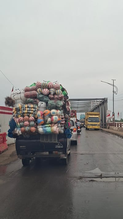 In Lhoknga, Indonesia, a heavily overloaded black ISUZU TURBO pickup truck is stopped in traffic on a wet road. The truck's bed is piled high with a large assortment of items, including many colorful, striped balls that appear to be volleyballs or similar sporting equipment, as well as numerous cylindrical objects wrapped in plastic and netting, and some white, stuffed-like figures. A silver car is to the right of the truck, and a motorcycle is in front of the car, also stopped in traffic. Further ahead, a yellow truck is visible. The scene appears to be on a bridge, indicated by the steel structure overhead. The sky is overcast. The road surface is wet and reflects the vehicles and sky. Red and white flags are visible on the right side of the bridge.