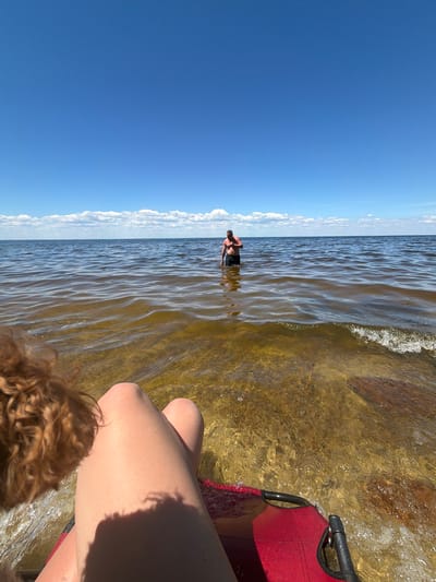 In Lebedivka, Ukraine, on a sunny day with a clear blue sky and scattered white clouds, a person is partially submerged in shallow, clear water. The water has a golden hue, and the sandy bottom is visible. The person is wearing dark shorts and has sunburn on their shoulders. In the foreground, a person's legs and lap are visible, sitting in a red chair. A dog's reddish-brown fur is also visible in the lower left corner of the frame. The water ripples and reflects the sunlight. In the distance, a thin line of clouds appears above the horizon where the sky meets the water.