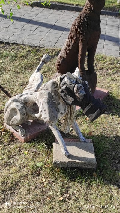 The image shows a wooden sculpture of a dog wearing a gas mask and a bandana, placed outdoors on a grassy area with some scattered leaves. Behind the dog sculpture, a portion of another dark wooden sculpture of a sitting animal, possibly a bear or a larger dog, is visible. The ground is covered with dry grass and some fallen leaves. In the background, a paved walkway with square tiles and a shadow cast by sunlight can be seen. The overall lighting suggests it is daytime. The image contains a watermark at the bottom left corner reading "REDMI NOTE 9 PRO AI QUAD CAMERA" and a timestamp at the bottom right corner reading "2026/7/8 18:36". The location in Iisalmi, Finland is not specified in the image itself, but this is a general description of the visual content.
