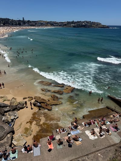 Aerial view of a crowded beach on a sunny day. Waves crash along the shoreline where numerous people swim. Others sunbathe on the sand and a concrete platform, while a few explore rocky areas. Buildings line the beach in the distance.