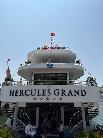 The image shows a vertical view of the Hercules Grand boat in Tuan Chau Ward, Vietnam. The boat has a multi-tiered white structure with colorful flags strung around its edges. "HERCULES GRAND" is printed vertically on a white surface with three gold stars. People can be seen near the entrance, including a person in a white shirt and black tie carrying a basket. A Vietnamese flag flies atop the boat. A sign displaying the registration number "99 QN-9456" is visible. In the background, the sky is clear blue.