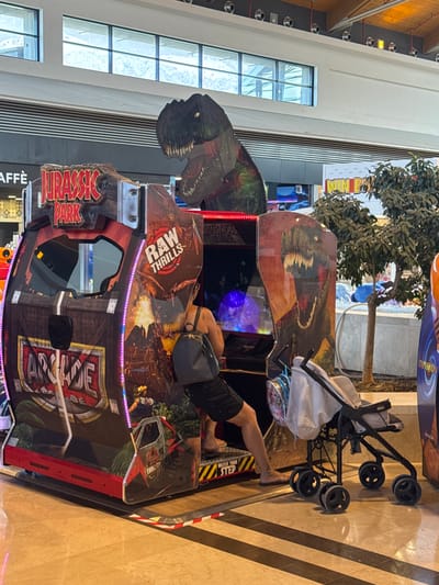 This media file features a Jurassic Park arcade game in what appears to be an indoor entertainment venue, possibly a mall, in Carini, Italy. The game itself is a large, upright cabinet with vibrant graphics depicting dinosaurs, including a T-Rex head prominently displayed on the side. The text "JURASSIC PARK" is clearly visible on the cabinet.
A person, whose legs and torso are visible, is lying on top of the arcade machine, facing away from the camera. They are wearing dark shorts and what appears to be a black backpack or bag is on their back. Their feet are bare, and they have a tattoo on one leg. The person's pose suggests they might be resting or have fallen asleep on the machine.
Next to the arcade game, a black baby stroller with a white bag inside is parked. The stroller is a compact model with four wheels.
The background shows architectural elements of an indoor space. On the left, there are large windows with a grid pattern, possibly looking out onto another part of the building or an outdoor area. The floor is a patterned tile with diagonal lines in beige and dark brown. In the upper background, there is a small, artificial tree with dark green foliage.
The lighting appears to be artificial, typical of an indoor environment, and there are no obvious cues indicating the time of day or weather conditions. The overall scene seems to capture a moment of rest or inactivity in a lively entertainment setting. The visible text includes "JURASSIC PARK" and "RAW THRILLS" on the arcade game, and potentially "TYPHOON" on a banner in the background. Another sign that appears to read "CAFFÈ" is visible at the bottom left.
