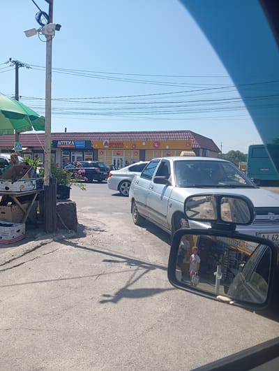 A daytime outdoor scene shows a street in Vasylkiv, Ukraine, with a focus on a silver sedan in the foreground. The car has a taxi sign on its roof and is parked on the right side of the frame. The vehicle's side mirror reflects a young girl holding a bag and walking with an adult. Behind the sedan, a turquoise bus is visible. Across the street, a row of storefronts includes an "Apteka" (pharmacy) and a "Magnit Multimarket." A security camera is mounted on a pole on the left side of the image, near a green umbrella and a market stall displaying boxes of bananas. The sky is clear and blue.