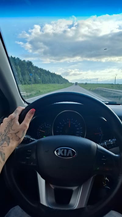 This is a photograph taken from inside a car, with the steering wheel and dashboard prominently featured. A woman's hand, with a pink painted nail and a tattoo on her forearm, is visible gripping the steering wheel. The car appears to be moving along a road, as indicated by the blur of the passing scenery and the visible speedometer on the dashboard which reads approximately 100 km/h. The road stretches ahead, flanked by lush green trees and fields, and the sky above is a bright blue with fluffy white clouds. The time of day seems to be daytime, likely late morning or afternoon, given the bright sunlight. The car is identified as a Kia by the logo on the steering wheel. The overall impression is one of a scenic drive on a pleasant day.