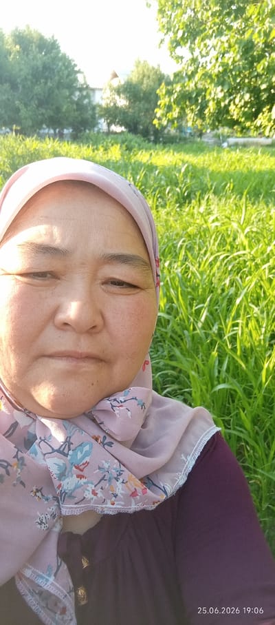 A close-up selfie is taken outdoors, featuring a woman wearing a pink hijab with a floral pattern and a dark purple dress. The woman's face is illuminated by the setting sun, casting a warm glow and highlighting some wrinkles. She has a neutral expression, looking directly at the camera.
The background is a vibrant field of tall green grass, bathed in the golden light of either sunrise or sunset. Behind the grass, trees are visible, some with dense foliage, and a hint of buildings or structures can be seen in the distance. The overall scene suggests a peaceful, natural setting.
The image contains visible text in the bottom right corner: "25.06.2026 19:06". This indicates the date and time the photograph was taken. Given the time and the lighting, it is likely late afternoon or early evening, and the weather appears to be clear. The location is identified as Chon-Kara, Kyrgyzstan.