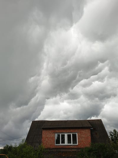 The image shows the roof and upper story of a brick house under a dramatic, cloudy sky. The sky is filled with heavy, gray storm clouds that obscure the sun. The house has a tiled roof and two windows. The lower portion of the image shows some greenery, likely trees and bushes. The scene appears to be in a rural or semi-rural setting, possibly in Medvedyovka, Russia, based on the context provided. There are no people or specific actions depicted. The overall mood is somber and indicative of approaching or ongoing inclement weather.