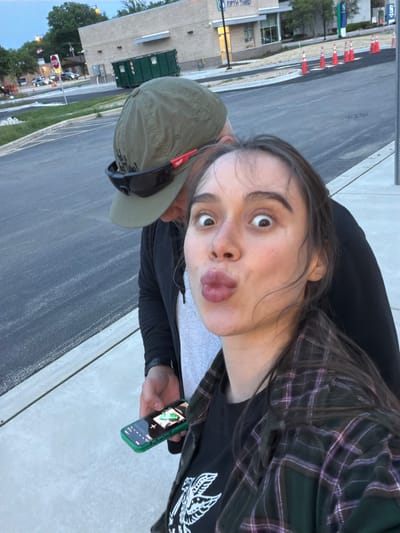 A selfie depicts a woman with light skin, brown hair, and light eyes making a duck-like pouting expression. She is wearing a black shirt, a green and purple plaid shirt, and a black jacket. She is holding a green-cased smartphone in her right hand. Beside her is a man wearing a green baseball cap with sunglasses attached to the bill. The background shows a street in Morton Grove, Illinois. A Fifth Third Bank building is visible in the background, as well as orange construction cones and other buildings. The road is asphalt, with a concrete sidewalk to the right of the people.
