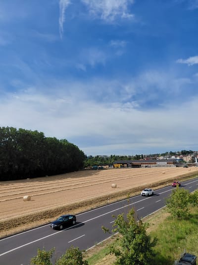 A horizontal shot captures a sunny day in Ecublens, Switzerland, with a clear blue sky dotted with white clouds. A highway runs diagonally across the lower portion of the image. On the highway, a black sedan is in the left lane, and further ahead, a white SUV and a red car are in the right lane. To the left of the highway, a field of harvested wheat is visible, with rows of straw bales scattered across it. A dense line of trees forms the backdrop to the field. In the distance, a town with buildings and a large warehouse is visible on a hill. The overall scene depicts a rural landscape on the outskirts of a city on a bright summer day.