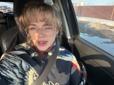 A young woman with light brown hair and gold-rimmed glasses is seated in the front passenger seat of a vehicle in Азьмушкино, Russia. She winks her right eye and extends her tongue slightly, looking directly at the camera. She wears a rust-colored high-necked top underneath a patterned jacket featuring dark blue, white, and multicolored abstract designs, secured by a black seatbelt. A small blue dangle earring is visible on her right ear. Strong sunlight illuminates her from the left side of the frame. Through the car's right-side window, an exterior scene reveals patches of melting snow and dark, wet ground adjacent to a corrugated reddish-brown metal fence. Beyond the fence, a light-colored building with dark vertical elements is visible under a clear, bright sky.