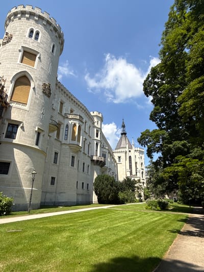 The image depicts a section of Hluboká Castle in Hluboká nad Vltavou, Czechia. The foreground features a meticulously manicured green lawn with visible mowing patterns, bordered by a paved pathway on the right side. A lamppost stands on the left side of the pathway, adjacent to the castle's wall.
The castle itself is a prominent subject, constructed of light-colored stone and characterized by a large circular tower on the left and a more elongated wing extending towards the right. Architectural details include Gothic-style windows, some with wooden shutters, decorative elements on the facade, and a balcony structure. Further in the background, another section of the castle is visible, featuring a distinctive conical tower with a spire.
Lush green trees with dense foliage flank the right side of the castle, creating a natural boundary. The sky above is a clear blue with scattered white clouds, suggesting a sunny day. The overall scene conveys a sense of grandeur and well-maintained historical architecture set within a park-like environment. There are no people or animals visible in the image.