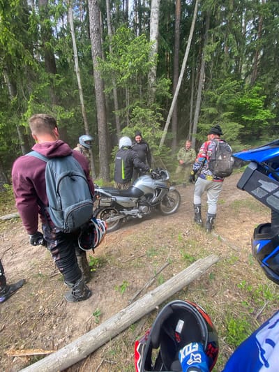 A group of people are gathered on a dirt path in a wooded area. The path is lined with tall trees. A motorcycle is parked on the path. Several people are standing near the motorcycle, while others are further down the path. A log lays near two motorcycle helmets, which are near a patch of green vegetation. The scene is located in the Парэцкі сельскі Савет, Belarus.