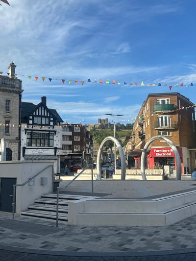 The image depicts a street scene in Dover, United Kingdom. In the foreground, there is a modern concrete plaza with a staircase leading up to it. The plaza is decorated with large, abstract, metallic archways. Beyond the plaza, buildings line the street. To the left, a light-colored stone building with arched windows is visible, adjacent to a black and white timber-framed building. In the center of the frame, a castle-like structure is visible on a hill in the distance, against a blue sky with scattered clouds. Colorful triangular flags are strung across the sky. Further down the street, to the right, a brick building with multiple balconies is seen. A red shopfront with the text "Furniture & Electrical" is prominent. Several cars are parked or driving on the street. A few individuals are present, one walking on the sidewalk and another sitting on a bench. The overall scene suggests a public space in a town with a historical landmark.