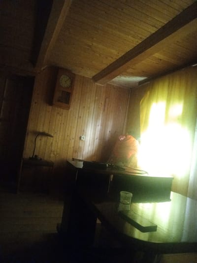 This media file captures an interior scene in a room with wooden paneling on the walls and ceiling. A prominent wooden beam runs across the ceiling. On the wall, a clock with a round face and Roman numerals is visible. Below the clock, a desk lamp with a dark, curved arm is positioned on a dark surface, likely a desk or table. A light switch is also mounted on the wall. To the right of the desk, there is a large window covered by a bright yellow curtain, through which sunlight is streaming, creating a strong glare. In front of the window, a large orange bag or pillow rests on a surface. A table with a dark, reflective surface is in the foreground, with a small clear plastic cup and a dark rectangular object on it. The room appears to be dimly lit, with the exception of the bright light from the window and the light from the lamp. The overall impression is one of a rustic or cabin-like setting. No people are visible in the image. The time of day is suggested to be daytime due to the sunlight streaming through the window. The location is identified as Kamayevo, Russia. There is no visible text in the media file.