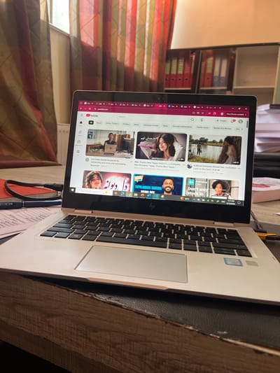 The image shows a laptop with the YouTube interface open, displaying various video thumbnails. The laptop is positioned on a wooden surface, possibly a desk or table, with a notebook and pen lying to the left. In the background, striped curtains are visible, and behind them, shelves filled with binders and books can be seen. The lighting suggests an indoor environment, with natural light coming from the left, casting shadows. The screen of the laptop shows a Google Chrome browser window with multiple tabs open, including YouTube, and other general browsing content. The overall scene depicts a workspace, likely in a home or office setting. The location in Kabul, Afghanistan is not discernible from the visual elements of the image.