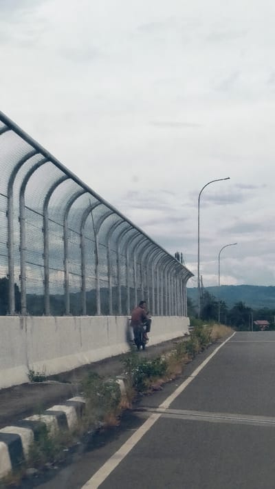 This daytime image, set in Lonbaroh, Indonesia, captures a stretch of road under an overcast sky. A prominent feature is a long, tall metal mesh fence with a curved top, likely a sound or safety barrier, extending diagonally across the left side of the frame. It rests on a concrete wall that separates the road from the unseen area beyond the barrier.
In the midground, a man wearing a reddish-brown shirt is seated on a dark motorcycle, parked alongside the concrete wall beneath the fence. He appears to be resting or waiting, looking forward. The road itself is asphalt with clear white lane markings, and a section of black and white striped concrete barriers line the inner edge of the road, partially overgrown with weeds and vegetation.
Further along the road on the right, two tall streetlights with curved arms stand against the light grey, cloudy sky. In the distant background, a range of green mountains is visible, with a few small buildings or houses nestled at their base. The overall impression is a somewhat desolate, functional roadside environment under diffused daylight. No text is visible in the image.