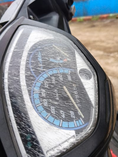 A close-up, angled view reveals the dashboard and gauge cluster of a black Yamaha scooter or motorcycle, positioned outdoors in Kyauk Hsoke, Myanmar. The primary subject is the speedometer and odometer, which are heavily obscured by a textured layer of dust, grime, or fine cracks on the protective plastic cover. The speedometer displays a range from 0 to 180 Km/h, with a prominent blue arc marking the 0 to approximately 100 Km/h interval. The speed needle rests at 0 Km/h. An odometer reading of "19865" is partially visible. A Yamaha logo is present on the right side of the gauge. The background, out of focus, shows a light brown, possibly unpaved, ground surface with a bright blue structure or wall and a horizontal orange line visible above it. No individuals or active movements are depicted.