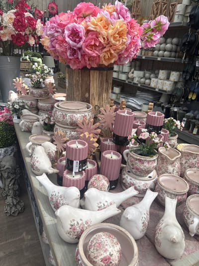 In Fritzlar, Germany, a retail display features a collection of floral-themed decorative items. The foreground is dominated by several ceramic bird figurines, each adorned with pink and white floral patterns. Adjacent to the birds are cylindrical pink candles with visible wicks, some stacked and others individually placed. Various ceramic pots and planters, also decorated with similar floral motifs, are arranged throughout the display. A large bouquet of artificial pink and orange roses is prominently featured in a wooden barrel towards the top of the frame. In the background, shelves display more ceramic items and other merchandise, suggesting an indoor retail environment. The overall impression is one of a curated display of decorative home goods, likely in a garden center or gift shop.