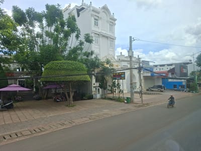 This image depicts a street scene in Liên Nghĩa, Vietnam, with a prominent white, multi-story building characterized by ornate architectural details and multiple windows. In front of this building, there is a paved walkway with several trees, including one with a meticulously trimmed, square canopy. Two purple umbrellas are set up on the walkway, and two motorcycles are parked nearby. To the right of the white building, a blue structure with the words "CUP COFFEE" is visible. A dark-colored SUV is parked in front of a building to the left of the coffee shop. A man wearing a red helmet is riding a motorcycle down the paved street in the foreground. The sky is overcast with scattered clouds.