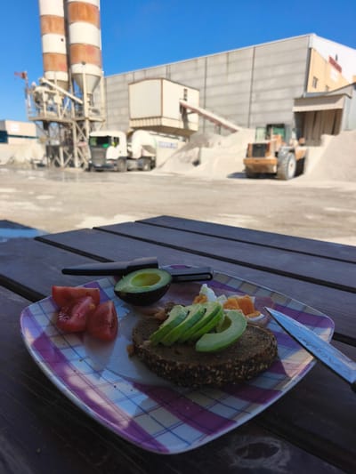 The image presents a plate of food on a wooden picnic table, with an industrial backdrop. The plate contains sliced tomatoes, half an avocado, sliced avocado on a piece of dark bread, and chopped eggs and ham. A knife and a peeler are also on the plate. The background shows a concrete plant or quarry, featuring silos, trucks, and heavy machinery. The scene is set in the city of Polígon Industrial Dels Vents, Spain, indicated by the type of industrial structures present. The sky is clear and blue, suggesting a sunny day. No people are visible.