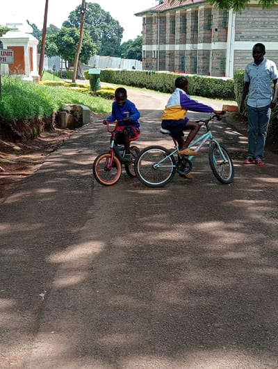 An outdoor scene captured on a sunny day in Riara Ridge, Kenya, shows three individuals on a paved road lined with green bushes and trees. In the foreground, two children are on bicycles. The child on the left, wearing a blue long-sleeved shirt and red plaid shorts, is seated on a small red bicycle with training wheels, looking towards the viewer with a cheerful expression. The second child, positioned slightly to the right, is on a larger light blue bicycle, wearing a multi-colored (yellow, white, purple) long-sleeved top and dark shorts, looking forward.
To the right of the children, an adult male stands on the side of the road, dressed in a light blue short-sleeved button-up shirt, dark trousers, and red shoes, observing the children. The paved road features dappled shadows, suggesting overhead trees and bright sunlight.
In the background, a large building with a stone or brick facade and a tiled roof is visible on the right. On the left, there's a white and orange entrance structure with a sign that partially reads "LIMIT" and "P/H", likely indicating a speed limit. Beyond the entrance structure, more greenery and another building structure with a corrugated metal fence are seen. A green trash can is also visible between the road and the background buildings. The overall atmosphere appears calm and leisurely.