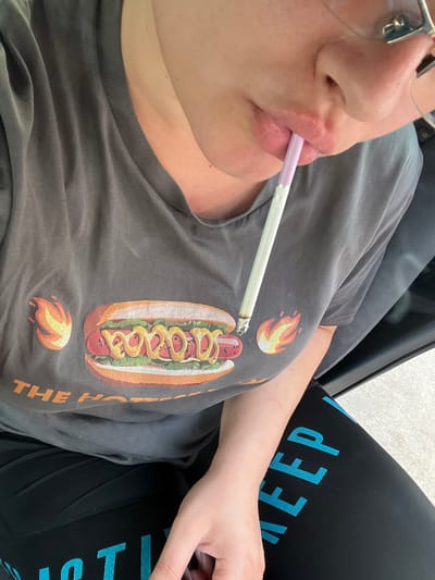 A person, partially visible from the chest up, is holding a lit cigarette with a pink filter between their lips. They are wearing a dark gray t-shirt with a graphic of a hot dog with flames on either side and the words "THE HOT..." visible below it, and black leggings with blue lettering that appears to say "...KEEP". The person is seated in what looks like a car. The background is blurred and out of focus, suggesting an indoor or shaded environment. There is no indication of the city of Gorni Dabnik, Bulgaria in the image.