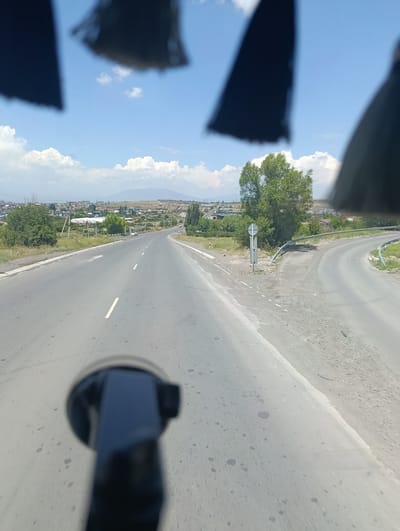 This media file depicts a view from inside a vehicle, likely a car, looking out onto a road in Jrvej, Armenia. The scene is set on a sunny day with scattered white clouds against a clear blue sky. The road ahead splits into two lanes, one going straight and another curving to the right. The road is paved and shows signs of wear, with some dark spots and cracks visible on its surface.

On either side of the road, there are grassy areas and patches of dirt. Trees with lush green foliage are scattered throughout the landscape, with a prominent cluster of trees to the right of the road. In the distance, there is a town or city with various buildings and structures, and further beyond, hills and mountains are visible. The overall impression is that of a rural or semi-urban environment during daytime.

A black object, possibly a car mount for a phone or camera, is prominently in the foreground, slightly out of focus. The tops of black car accessories or parts of the vehicle's interior are also visible in the blurred upper portion of the image, framing the sky and distant landscape. No people are clearly visible in the image. The weather appears warm and pleasant, suitable for travel. No visible text is present in the image.