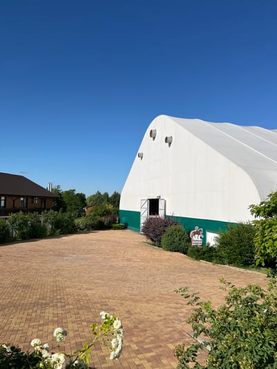 The image captures a sunlit outdoor scene at a facility in Plastunivska, Russia, under a clear blue sky. In the foreground, a paved area made of interlocking reddish-brown bricks leads towards a large, white, arched tent-like structure. This structure, with a green band around its base, has an open doorway. To the left of the paved area, a two-story wooden house with a dark brown roof is visible, surrounded by lush green trees and bushes. A sign with a silhouette of a horse and rider, and text in Russian, is placed in front of the tent. The middle ground and background are filled with dense green foliage, indicating a natural or park-like environment. White flowers are blooming in the lower left corner of the frame.