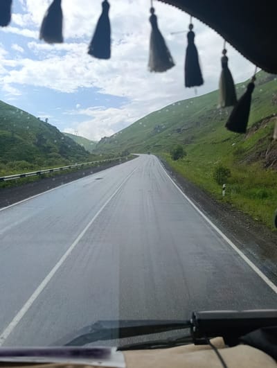 The image depicts a view from inside a vehicle, looking out at a wet, paved road winding through a valley. The road is flanked by steep, green, grassy hillsides. Overhead, the sky is partially cloudy with patches of blue. Several dark tassels hang from the top of the frame, obscuring part of the sky and the upper reaches of the hills.

The road appears to have been recently rained upon, with reflections visible on its surface. A metal guardrail runs along the left side of the road, and on the right, a white and red striped marker post is visible near the edge of the pavement. The hills are covered in lush green grass, with a few scattered trees and rocks. In the distance, a large metal structure, possibly a power line tower, is visible on one of the hills. The weather appears overcast with intermittent rain, suggested by the wet road and the droplets visible on the windshield. The time of day is indeterminate, but the lighting suggests daytime. The overall mood is one of travel through a scenic, somewhat damp, natural landscape. No people are visible in the image.