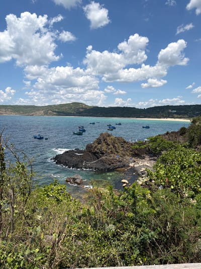 A bright, sunny day with a blue sky filled with puffy white clouds is captured in this medium-angle shot of a coastal landscape. The foreground is dominated by lush green foliage and shrubbery, some of which is prickly, obscuring a wooden railing. Beyond the vegetation, a choppy, deep blue ocean stretches out, dotted with several small, blue fishing boats. To the right, a rocky outcrop of dark, basaltic columns juts into the sea, with a few people visible on its upper surface. The middle ground features a white sandy beach backed by rolling, green hills that extend into the distance.