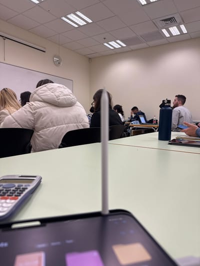 The image depicts an indoor classroom or lecture hall in Kfar Monash, Israel, taken from the perspective of a student's desk. In the foreground, a light green desk surface holds a dark-cased tablet, likely an iPad, with a white Apple Pencil magnetically attached to its side. A silver and black scientific calculator is also partially visible on the desk to the right.

The scene features several students seated at desks, facing away from the camera. The closest student wears a prominent white puffer jacket and has light-colored hair. In front of them, another student in dark clothing is visible, head slightly bowed, seemingly engaged with a task. Further down the row, a man with a beard, wearing a light grey hoodie, is looking at a small electronic device. A blue water bottle rests on the desk beside him. Other students are visible further in the background, some with open laptops on their desks, indicating an environment of study or lecture.

The room has light-colored walls and a ceiling equipped with multiple rectangular fluorescent light fixtures and air vents. A large whiteboard is mounted on the left wall, and above it, a circular clock displays the time as approximately 10:20 or 10:25. The atmosphere is one of focused academic activity. No distinct text is legible on the tablet screen, though the calculator's buttons are visible. The overall impression is a contemporary educational setting where students utilize both digital and analog tools.