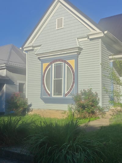 The image shows the side of a light blue house with a unique circular window. The window is framed with dark blue and yellow accents, and the glass is divided into sections with a circular motif. A portion of a neighboring house with a gray metal roof is visible to the left. Green bushes and tall grasses are in the foreground, with a stone foundation visible at the base of the house. The sky is clear and blue, suggesting a sunny day. The scene appears to be residential. There is no visible text or people in the image.