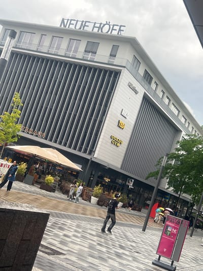 This image depicts an outdoor urban scene in Herne, Germany, specifically featuring the "Neue Höfe" complex and the surrounding plaza, with elements suggesting proximity to the Rathaus. The weather is overcast.
The central subject is a multi-story, modern building with "NEUE HÖFE" prominently displayed in large letters on its roofline. The building facade is a combination of white and grey panels, featuring distinct sections of vertical grey fins that extend across multiple floors. Below the "NEUE HÖFE" sign, a row of windows with balconies and dark blinds is visible. On the lower part of the building's facade, multiple signs indicate businesses: "FalkoGroup", "NKO", "TED", and "GOLD'S GYM". Further to the left, a sign partially visible spells "RATHAUS".
At ground level, a wide, patterned plaza extends into the foreground, paved with alternating grey and white rectangular blocks arranged in a chevron or herringbone pattern. In the mid-ground to the left, an outdoor seating area of a restaurant or cafe is present, furnished with numerous tables and chairs, protected by a large, light-beige umbrella. Planters containing small green trees and shrubs are placed around this area. White and red plastic barriers are visible near the outdoor seating.
Several individuals are present in the scene. In the center foreground, a man wearing a black t-shirt, dark jeans, and white over-ear headphones walks towards the right, holding a white object, possibly a smartphone. To his left, a woman in a dark blue jacket and black pants walks away from the camera. Further back on the right side of the plaza, two women walk together; one wears a light grey top and jeans, the other a white top and jeans. Near a pink informational stand in the mid-ground, a group of people is seen, including individuals holding colorful umbrellas (one rainbow-striped, one solid pink), suggesting recent or ongoing rain. The pink sign, part of a "MAGENTA FAN-DEAL" advertisement, features a 'T' logo, indicating Telekom. Numerous other indistinct figures are scattered throughout the plaza and within the outdoor cafe area.
The environment includes several dark street poles and a large, dark, blocky structure in the far left foreground. Green trees are interspersed, adding natural elements to the urban setting. The sky is uniformly cloudy.