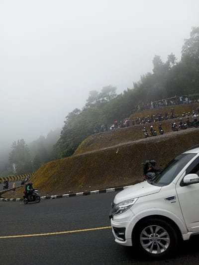 The image depicts a mountainous road scene in Phoxay, Laos, under conditions of dense fog and mist. A dark asphalt road, appearing wet, runs through the foreground and midground, featuring a solid yellow centerline and black and white striped curbs along its edge. On the far right, the front right quarter of a white SUV is visible, showing its headlight, bumper, and wheel. In the midground, a dark motorcycle with two riders wearing green jackets and helmets moves left on the road, with luggage secured to the rear. Additional motorcycles are visible further along the road.

To the right of the road, a multi-tiered, terraced hillside rises, covered with green and brown vegetation. Numerous individuals are congregated on these terraces, standing and sitting. A significant number of motorcycles are parked in rows across the various levels of the slope. One individual in dark clothing is observed near the base of the hillside, adjacent to the white SUV, leaning on a parked motorbike. The upper portions of the hills are lined with trees, largely obscured by the pervasive fog that dominates the upper half of the image. A yellow and black striped barrier is visible on a distant curve of the road to the left.