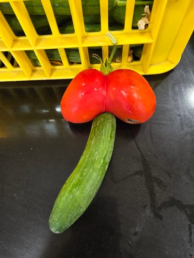 A close-up, top-down shot depicts a red tomato with a heart-like shape, resting on a dark, reflective surface. The tomato is positioned above a green cucumber, which is angled downwards. In the upper portion of the image, a yellow plastic basket is visible, containing what appear to be more cucumbers. The scene is indoors, suggesting a market or kitchen setting. The lighting is bright, casting reflections on the dark surface. There are no people visible in the image, and no context is provided about its location in Be'er Sheva, Israel.