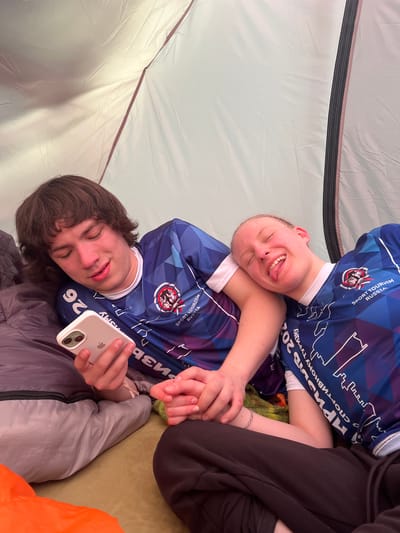 Two individuals wearing blue "Sport Tourism Russia" shirts are lying inside a tent. One person is holding a white iPhone. The shirt on the individual holding the phone has the number "26" printed on it. The other person has their tongue sticking out. The tent's interior is light gray. The setting is presumed to be in or around Lesnoye, Russia, based on the text on the clothing.