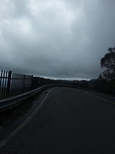 The image depicts an unpopulated section of a two-lane asphalt road in Bangtsho, Bhutan, under an overcast sky. The road, which appears wet, curves sharply to the right from the foreground towards the mid-ground. A solid white line delineates the left edge of the road, and a faded yellow dashed line is visible in the right lane, indicating the approximate center. To the immediate left of the road, a silver-grey metallic crash barrier runs along the curve. Behind this guardrail, a dark, spiked fence composed of numerous vertical slats extends along the periphery, suggesting a protective barrier overlooking a lower elevation or a precipice obscured by distance and atmospheric conditions. The sky is uniformly covered with dark grey clouds. On the far right, dark green foliage from a tree is visible, contrasting with the sky. The distant background shows a hazy outline of a landscape or structures, partially obscured by the low-hanging clouds or fog. No individuals, vehicles, or active interactions are present.