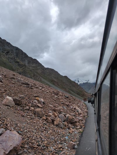 This is a photograph taken from inside a moving vehicle, looking out at a mountain road. The perspective is from the right side of the frame, with the side of the vehicle and its windows dominating the foreground and right edge. The road curves to the left, leading into a valley. On the left side of the road, a steep, rocky mountainside rises, covered in loose stones and scree. Some sparse green vegetation is visible on the higher slopes.
In the distance, the road continues into a mountain pass, where two trucks are visible. Further beyond the trucks, snow-capped mountains rise against a cloudy, overcast sky. The sky is filled with dramatic, grey clouds, suggesting inclement weather. The overall impression is one of a remote, rugged mountain landscape. This scene is located on the Baralacha Pass road, a high mountain pass in the Zanskar range, on the Leh-Manali Highway, in the region of Zing Zing Bar, India.