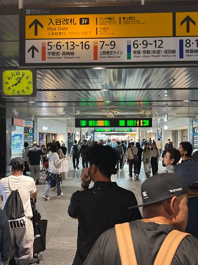 This forensic description details a bustling train station concourse at Ueno Station in Taito, Japan. Overhead, multiple signs direct passengers: a prominent yellow sign indicates the "Iriya Gate" on the 3rd Floor, along with "Panda Bridge Exit" and "Higashi-Ueno Exit." Below, a white sign delineates train lines and platform numbers: platforms 5, 6, 13-16 for the Utsunomiya-Takasaki Line; platforms 7-9 for the Ueno-Tokyo Line; and platforms 6, 9-12 for the Joban, Joban (Rapid), and Narita Lines. A square analog clock with a yellow face, displaying the JR logo, shows the time as approximately 11:25.

The concourse floor consists of light-colored tiles. Numerous individuals navigate the space, walking in various directions. In the immediate foreground, the back of a man wearing a white t-shirt, light-colored pants, and a black baseball cap is visible, carrying a black PUMA drawstring backpack and pulling a black tote bag on wheels. To his right, another man is seen from behind and the side, wearing a black t-shirt, a gold chain, and a backward black baseball cap, with his right hand near his ear.

Further into the scene, a diverse crowd of people, casually dressed, moves through the area. Several individuals are carrying bags or luggage. In the midground, a woman in a white hoodie over a plaid shirt walks away from the camera, pulling a white wheeled shopping bag. Green-on-black digital information screens are visible in the background, displaying train departure times and destinations, such as "20:11 UTSUNOMIYA" and "20:22 TAKASAKI" for platform 5. The ceiling features a grid pattern with integrated lighting.