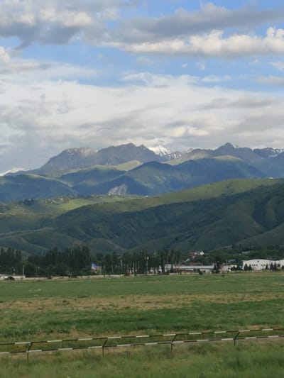 This media file captures a vast, scenic landscape in Қырғауылды, Kazakhstan. The scene is dominated by a series of rolling, verdant hills that recede into the distance, culminating in majestic, snow-capped mountains under a cloudy sky. The time of day appears to be late afternoon or early evening, with soft, diffused light casting gentle shadows across the terrain.
In the mid-ground, nestled within the hills, are scattered settlements with buildings and houses, suggesting a rural or semi-rural community. A line of tall, slender trees stands out along the lower part of the hills, adding depth and texture to the scene. The foreground features a wide, open field of green grass, with a striped barrier – likely a fence or a guardrail – running horizontally across the bottom of the frame. The grass in the foreground is a mix of vibrant green and drier, yellowed patches, indicating natural growth.
The weather is partly cloudy, with large, soft clouds interspersed with patches of blue sky. The lighting suggests a calm, possibly humid day, typical of mountainous regions. There are no people or animals prominently visible, focusing the viewer's attention on the natural beauty and the expansive panorama. The overall impression is one of tranquility and the grandeur of nature. No text is visible within the media file.