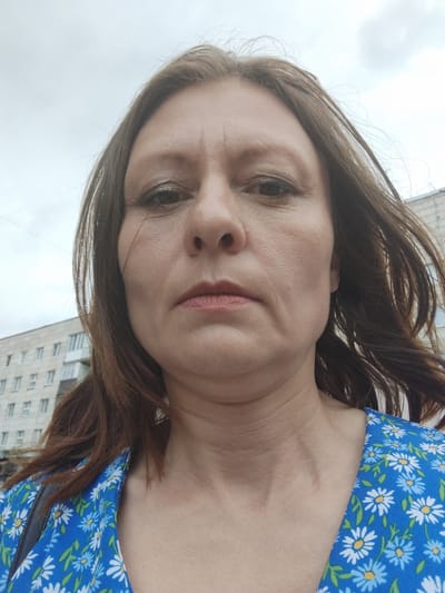This is a medium-shot, front-facing selfie of a woman taken outdoors. She has medium-length, wavy brown hair and appears to be in her late 30s or early 40s. She is wearing a blue dress with a white and blue floral pattern. The background shows a building that appears to be an apartment block, typical of Soviet-era architecture, suggesting an urban or suburban setting. The sky is overcast, indicating a cloudy day. The woman's expression is neutral. The lighting suggests it is daytime. There is no visible text in the image. Given the location context of Kirovsk, Russia, the building style and the overcast sky are consistent with this setting.