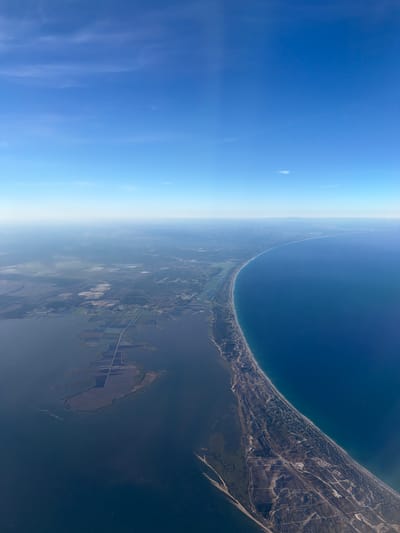 This high-altitude aerial image captures a sweeping view of a coastline under a clear, bright blue sky. The curved horizon suggests a wide-angle perspective from a significant elevation. The sky transitions from a deep blue overhead to a lighter, hazy blue-white closer to the horizon, indicating atmospheric depth.
The main geographical features include a vast expanse of deep blue ocean on one side, meeting a long, narrow sandy coastline. This coastal strip appears to be a peninsula or sand spit, separating the open ocean from a large, calmer body of water, likely an estuary or lagoon, on the landward side. Along the coastline, patches of vegetation, possibly dunes and low-lying forests, are visible, interspersed with signs of human development like roads and scattered buildings.
Further inland, the landscape reveals a mix of forested areas, cleared land, and agricultural fields. Distinctive circular irrigation patterns can be observed in some of the cultivated areas, indicating farming activity. Other structures and linear features, such as roads, crisscross the terrain. The overall lighting suggests it is daytime, under clear weather conditions. There are no people, explicit activities, or visible text in the image. The coastal morphology and agricultural patterns are consistent with the region of Grândola, Portugal.
