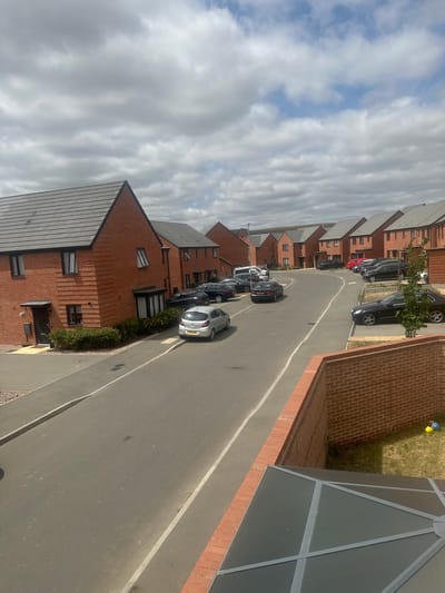 The image shows a view of a residential street in Wellingborough, United Kingdom, on a cloudy day. The street is lined with red brick houses with grey tiled roofs. Several cars are parked on the street and on driveways. A silver hatchback is parked on the left side of the street, and a black sedan is parked on the right. In the foreground, a brick wall and a glass-paneled roof structure are visible, suggesting a view from an elevated position, possibly a balcony or upper floor window. There are also two children's toys on the grass next to the brick wall, a yellow ball and a blue object. The sky is partly cloudy with patches of blue sky visible.