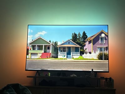 The scene is situated in a residential interior located in Straldzha, Bulgaria. A large, wall-mounted or stand-supported flat-screen television is the central focus, displaying a vibrant, animated street scene. The screen depicts three distinct, single-story houses with varied color schemes: a green house with red steps and a white garage on the left, a blue house with white steps and porch railings in the center, and a purple house with yellow trim and a white picket fence on the right. A paved street with a white broken centerline runs across the foreground of the screen, bordered by sidewalks and manicured green lawns. The sky above the houses is bright blue with scattered white clouds.
The wall immediately surrounding the television is illuminated by dynamic ambient lighting. The top section of the wall glows in shades of pale green and turquoise, consistent with the sky presented on the screen. The left and right side sections of the wall exhibit an orange-amber hue. Below the television, a dark, horizontal media console or shelf is visible. Resting on this surface, partially obscured by shadows, are several objects: a pair of reflective athletic sunglasses positioned near the left support of the television stand, and two clear glass perfume bottles with dark stoppers situated on the right side of the console. In the extreme lower-left foreground, a dark, textured fabric object, possibly part of a blanket or furniture, is visible. No individuals are present in the image, and no actions or interactions are depicted.