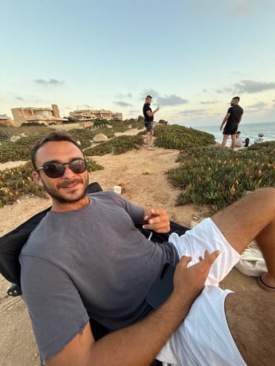 A man wearing sunglasses lies on the ground, casually dressed in a grey t-shirt and white shorts. A smartphone rests on his torso. The scene is outdoors, likely near a beach, with other people visible in the background, some near the shoreline. The landscape includes low-lying vegetation along a sandy area and buildings in the distance. The sky is visible and suggests it is either early morning or late evening.