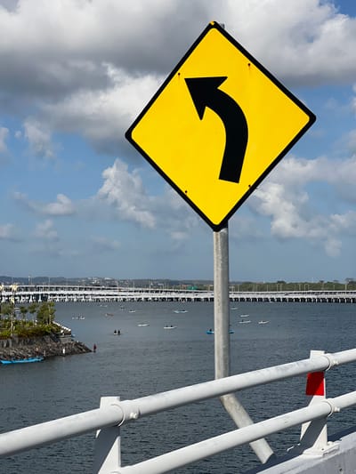 A yellow, diamond-shaped traffic sign with a black arrow indicating a left curve is prominently displayed in the foreground, mounted on a metal pole. The sign is positioned against a backdrop of a partly cloudy sky. In the middle ground, a body of water, likely the Badung Strait, separates the foreground from a long bridge or causeway that spans across it. The bridge appears to be a multi-lane structure with white railings and numerous lampposts. Numerous small boats, possibly kayaks or paddleboards, are scattered across the water, some with people in them. On the left side of the water, a small island or landmass with trees and a stone embankment is visible, with a few people standing near the water's edge. In the distance, the coastline is visible with more structures and greenery. This scene is likely located in South Denpasar, Bali, Indonesia, given the presence of the long bridge over the water which is a common feature in that area.