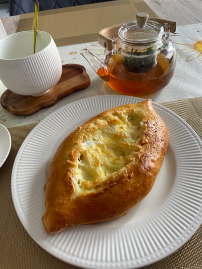 A golden-brown, boat-shaped pastry filled with melted cheese sits on a white, ribbed plate. Beside the plate, a clear glass teapot contains amber-colored tea and a mesh infuser with loose tea leaves. A white, ribbed mug with a gold spoon inside is positioned behind the teapot. The items are arranged on a neutral-colored placemat and a patterned tablecloth. The background shows a hint of a wooden table and a dark gray upholstered chair. The scene appears to be a casual meal, likely at a cafe or restaurant in Alzey, Germany.
